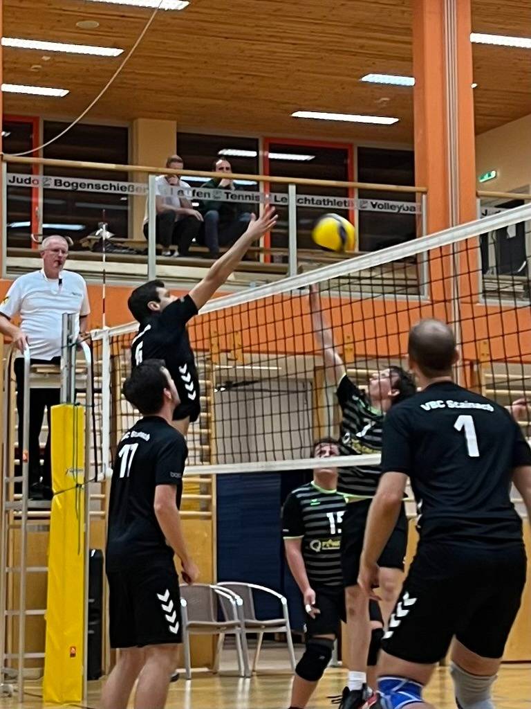 Blockduell am Netz: Spieler des VBC Stainach im schwarzen Trikot blocken einen Angriff, Schiedsrichter auf dem Hochstuhl daneben